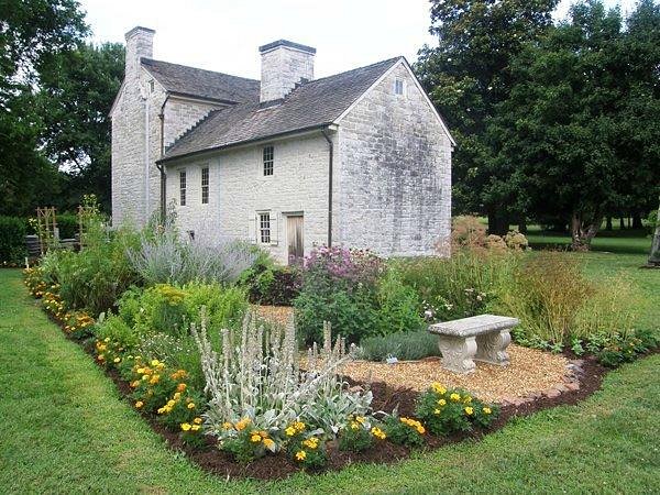 Historic stone house with lush flower garden, gravel path, and stone bench, surrounded by green lawn and trees. Perfect example of traditional cottage landscaping and architecture. Rock Castle, Hendersonville TN