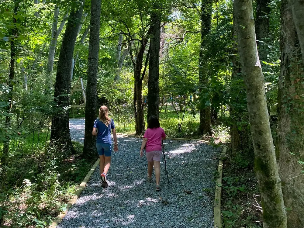 Two people walking on a gravel path through a wooded forest trail in daylight, surrounded by tall green trees and nature. Rock Castle, Hendersonville TN