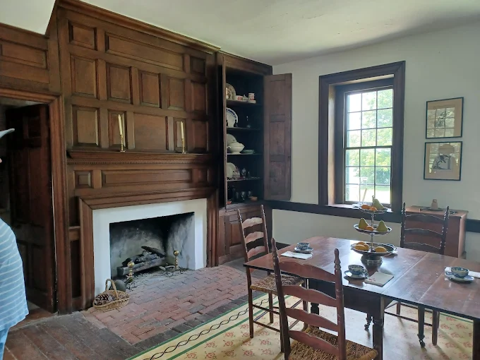 Historic dining room with wooden paneling, fireplace, antique chairs, and a window overlooking greenery; decorated shelves and a central table create a traditional, elegant atmosphere. Rock Castle, Hendersonville TN