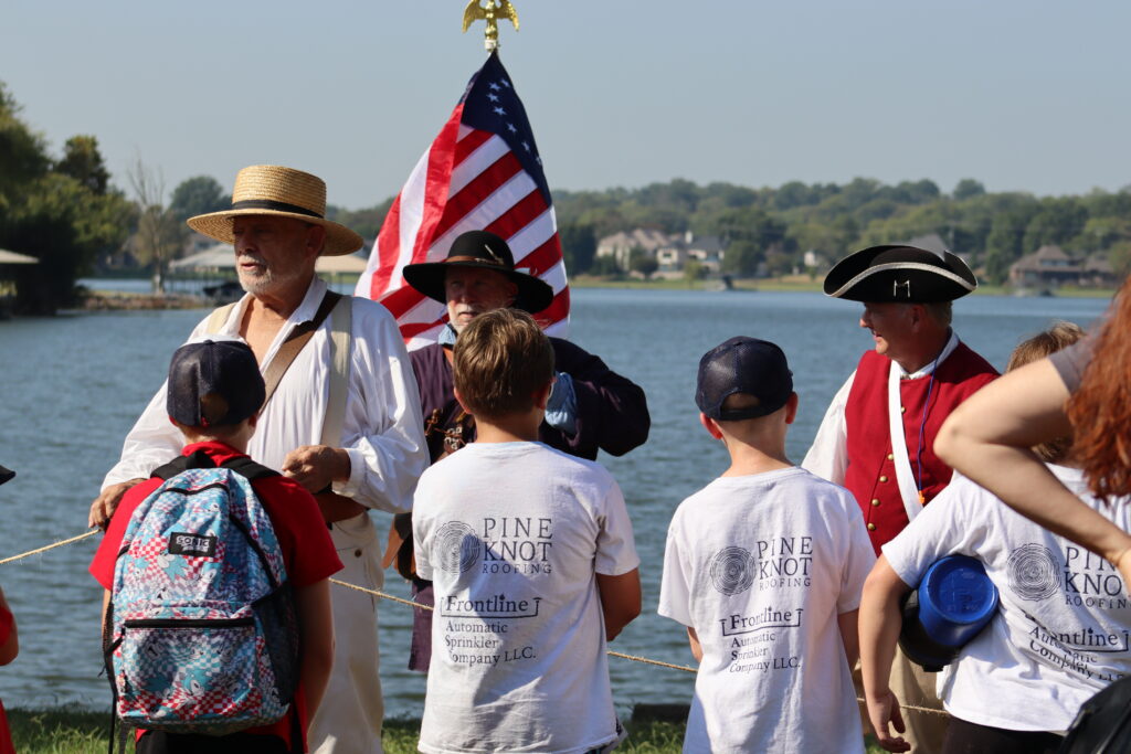 Adult male dressed in historical clothing of the late 1700-early 1800's talking with school aged boys. American flag waves behind the men.