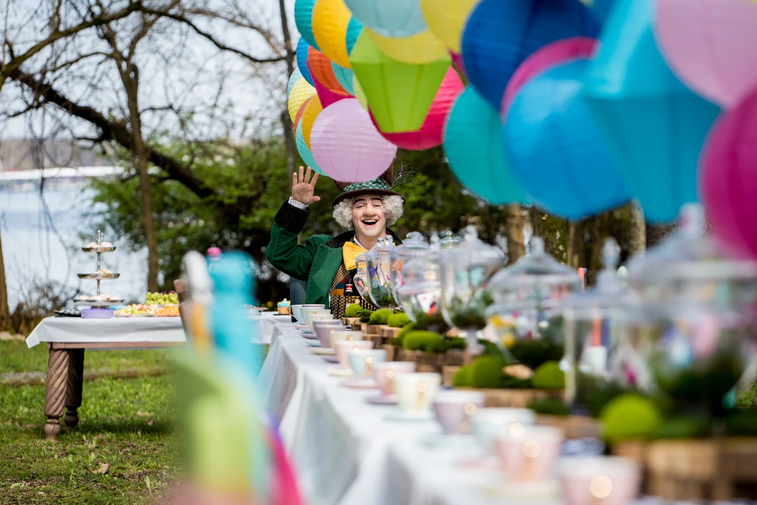Wonderland Egg Hunt themed tea party outdoors featuring the Mad Hatter waving, decorated with colorful paper lanterns hanging from trees at Historic Rock Castle.